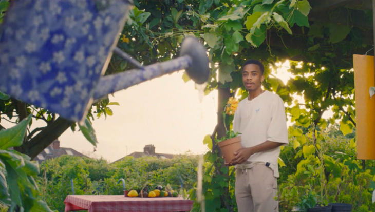A young person holding a yellow potted plant looking at a watering can.