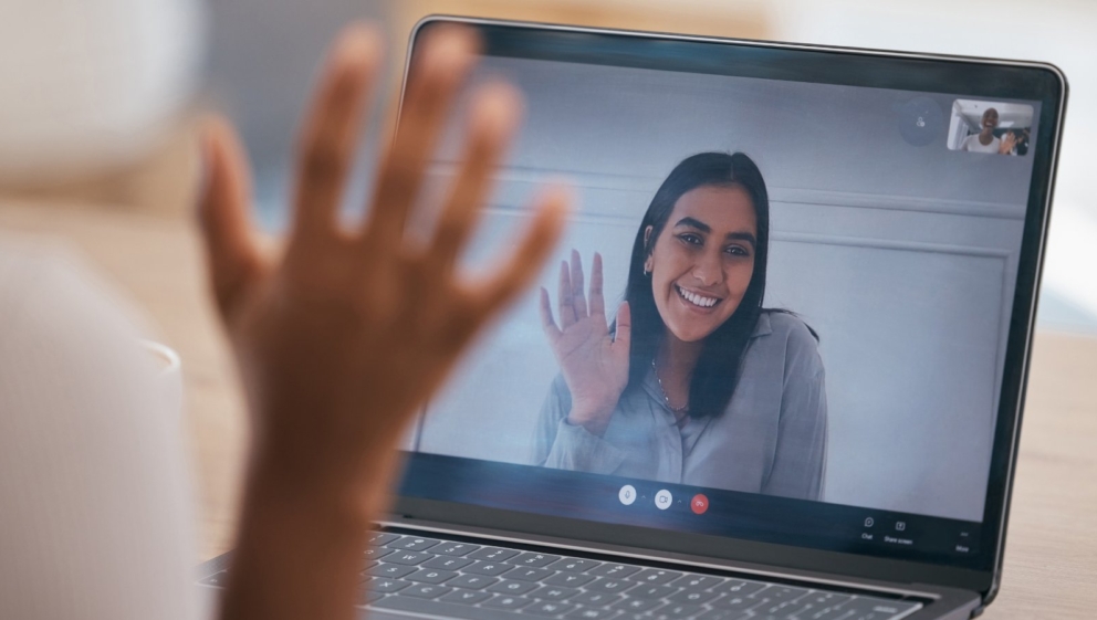 A person, on a laptop video call, waving to a person behind the laptop.
