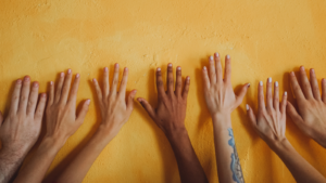 Six hands, in different shades and sizes, placed on a yellow wall.