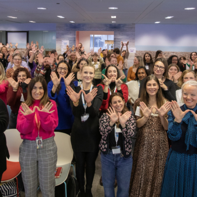 A large group of people at Women's Voices 2023, stood signalling the letter 'W' with their hands in celebration of International Women's Day