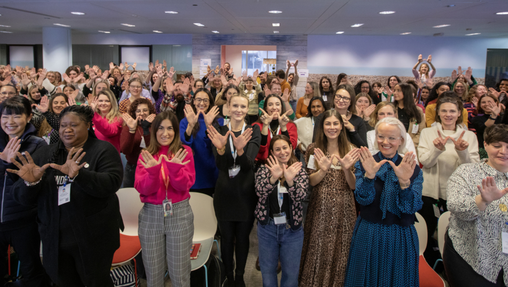A large group of people at Women's Voices 2023, stood signalling the letter 'W' with their hands in celebration of International Women's Day