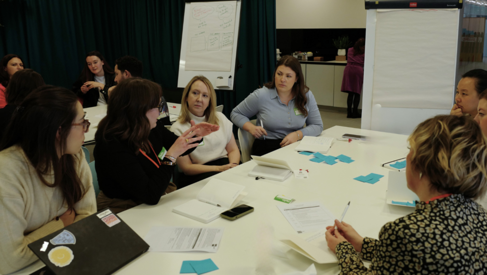 Two tables, with people sat around them talking. The tables are full of notes, and a whiteboard stands next to them.