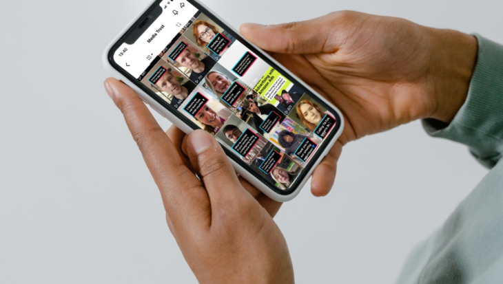 White background with a focus on person's hands holding a smartphone with the Media Trust TikTok account on the screen.