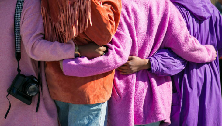 Four people are standing side by side with their arms around each other. They are wearing colorful clothing: a pink coat, an orange fringed jacket, a magenta coat, and a purple coat. A camera hangs from the shoulder of the person on the left. Their faces are not visible.