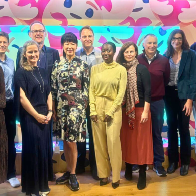 A group of men and women smiling at the camera. They are standing against a background of bright pink and blue dots.