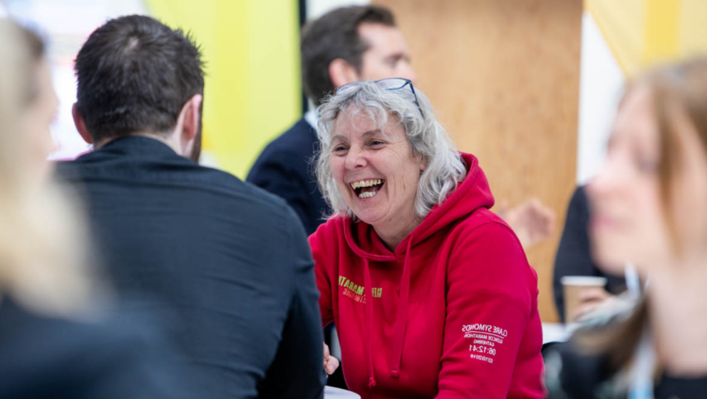 Two people sat in a busy room, chatting and smiling.