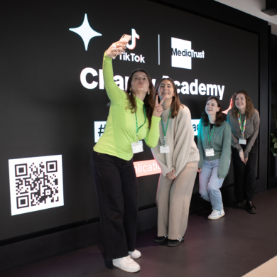 Five people posing for a selfie. They are stood in front of a large screen showing logos for TikTok, Media Trust and the TikTok Charity Academy