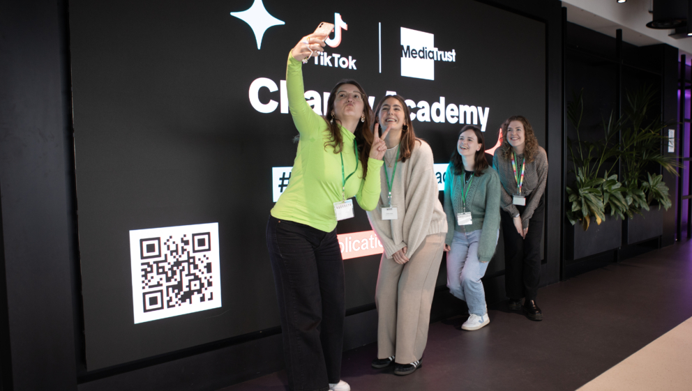 Five people posing for a selfie. They are stood in front of a large screen showing logos for TikTok, Media Trust and the TikTok Charity Academy