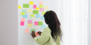 A black woman with long dark brown hair stands facing a white wall with her back to the camera. On the wall there are several green, pink, blue and orange post it notes.