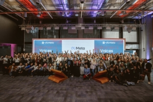 A group photo of over 100 people in a large event room. There is a large screen at the back, which reads 'women's voices 2025' and features both Meta and Media Trust's logos. The participants are waving their hands in the air, and are all gathered closely in front of the screen.