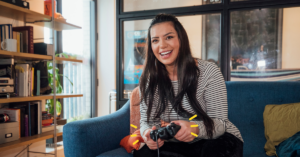 A woman with long dark brown hair is smiling into the camera with a game control in her hands. She is sat on a velvet blue sofa in what appears to be her home, playing games. She is wearing a grey and black long sleeve top.