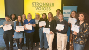 12 people posing with their certificates at the Stronger Voices graduation in 2025. Behind them is a Media Trust banner and a screen showing the Stronger Voices logo