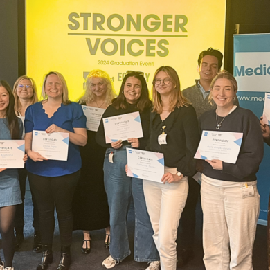 12 people posing with their certificates at the Stronger Voices graduation in 2025. Behind them is a Media Trust banner and a screen showing the Stronger Voices logo