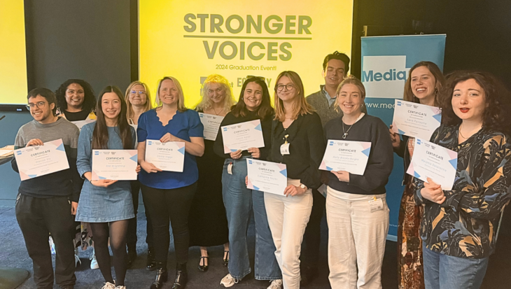 12 people posing with their certificates at the Stronger Voices graduation in 2025. Behind them is a Media Trust banner and a screen showing the Stronger Voices logo