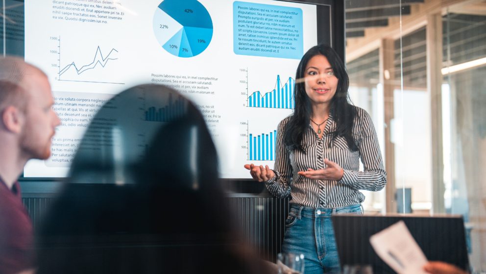 A person, in a meeting, stood presenting a screen with data and stats on it