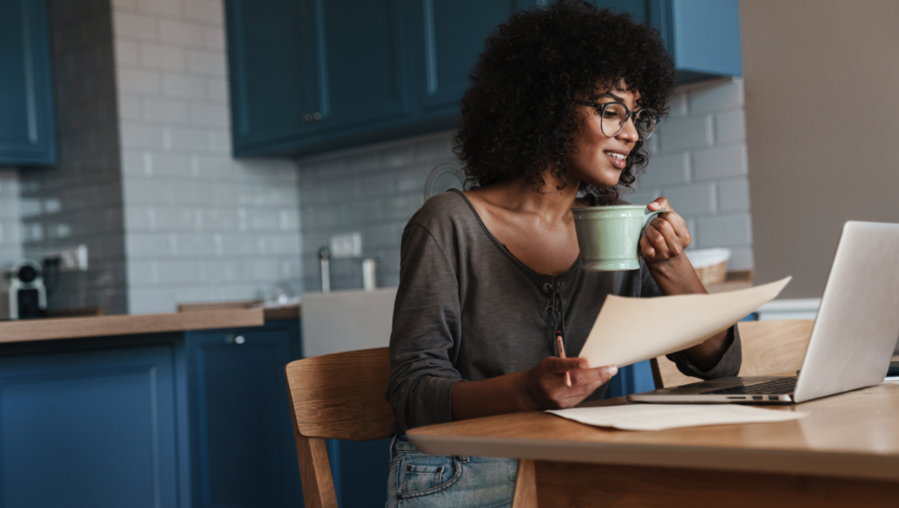 A person sat at their laptop, holding notes and a mug