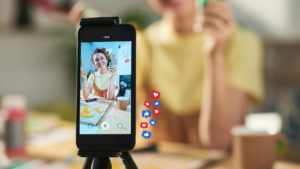 A phone propped up on a tripod sits at the front of the image. The screen shows woman with short brown hair is smiling and holding a green paint pot and paint brush. Behind the phone, you can see the blurred outline of the woman, alongside some blue and red 'like' and 'love' emojis.