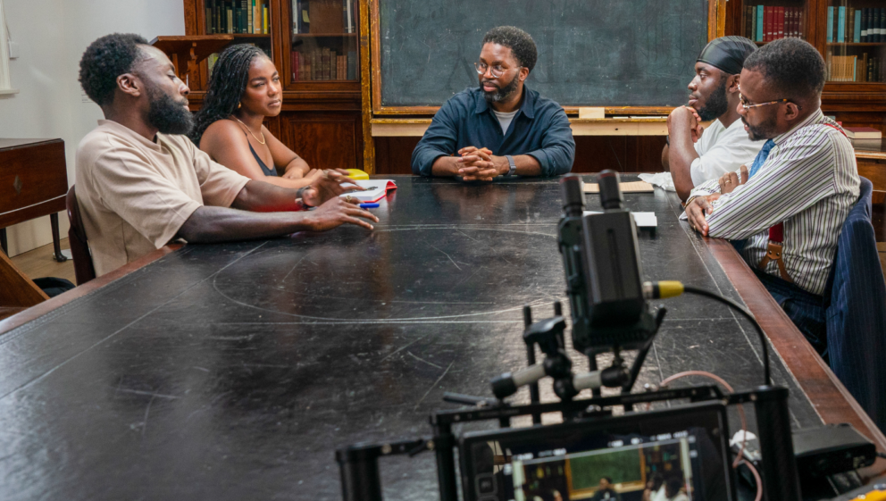 A BTS from the BBU film mandem, showing 5 people sat around a table, in front of a blackboard and bookshelves