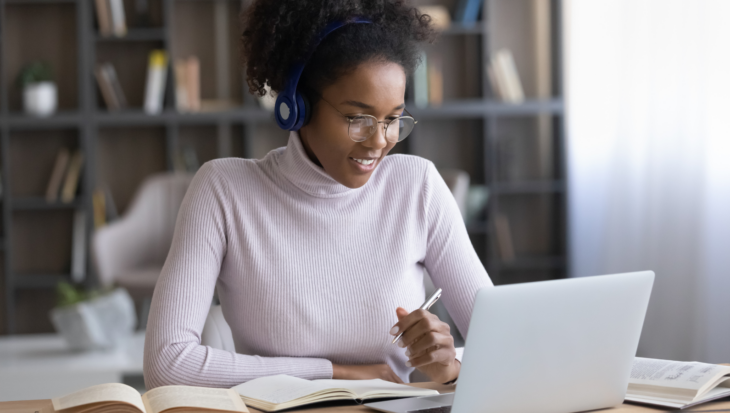 A person, wearing wireless headphones, smiling at their laptop and holding a pen