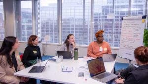 A group of women sit at a white table looking at a flipchart in a meeting room with their laptops open.