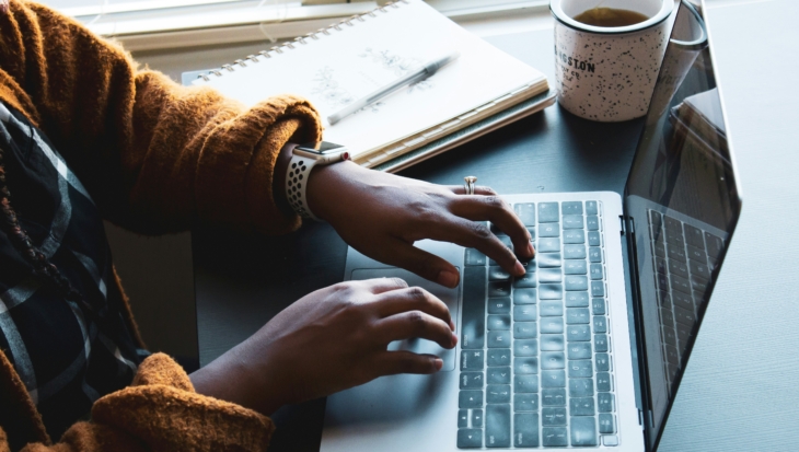A person with an orange jumper on is typing on their silver laptop. Next to the laptop is a notebook and mug.