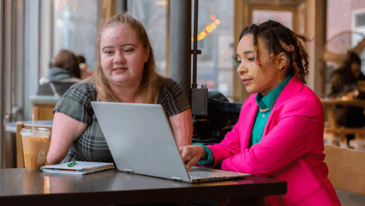 Two people who appear to be women sit side by side in a cafe in front of a laptop.