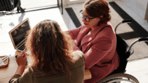 Two people sat, one in a wheelchair, at a desk and looking a laptop