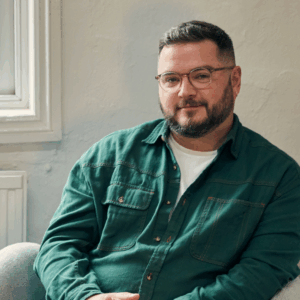 Colin is a man with short brown hair smiling softly at the camera. He is wearing a teal green shirt and has thin rimmed glasses.
