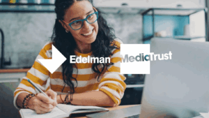A woman wearing blue rimmed glasses and a yellow and white striped top is sat smiling at her laptop. She has long black curly hair, and is writing in her notebook. Edelman and Media Trust logos