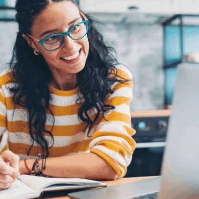 A woman wearing blue rimmed glasses and a yellow and white striped top is sat smiling at her laptop. She has long black curly hair, and is writing in her notebook.