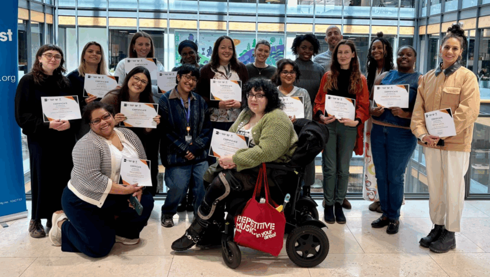 A group of individuals, holding Stronger Voices certificates, smiling in an office lobby with large windows in the background