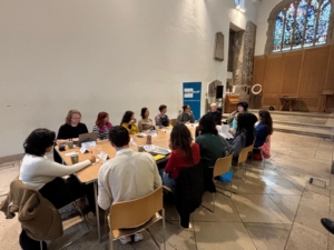 A group of people sat around a table in a church building, with a large stained glass window behind