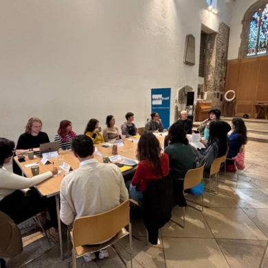 A group of people sat around a table in a church building, with a large stained glass window behind