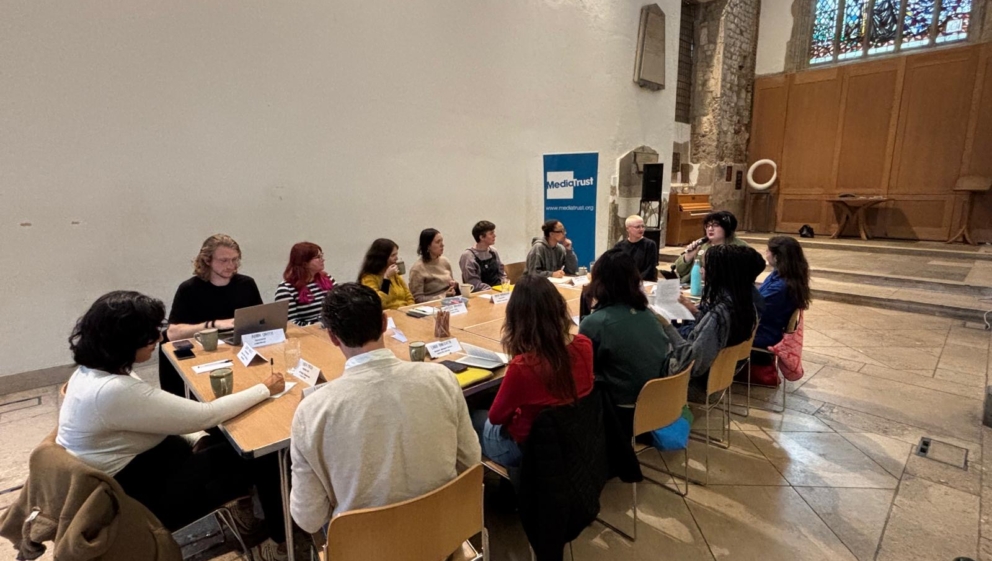 A group of people sat around a table in a church building, with a large stained glass window behind