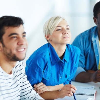Four people, sat smiling as they listen to a session. They have notes, pens and a coffee cup on the table in front of them.
