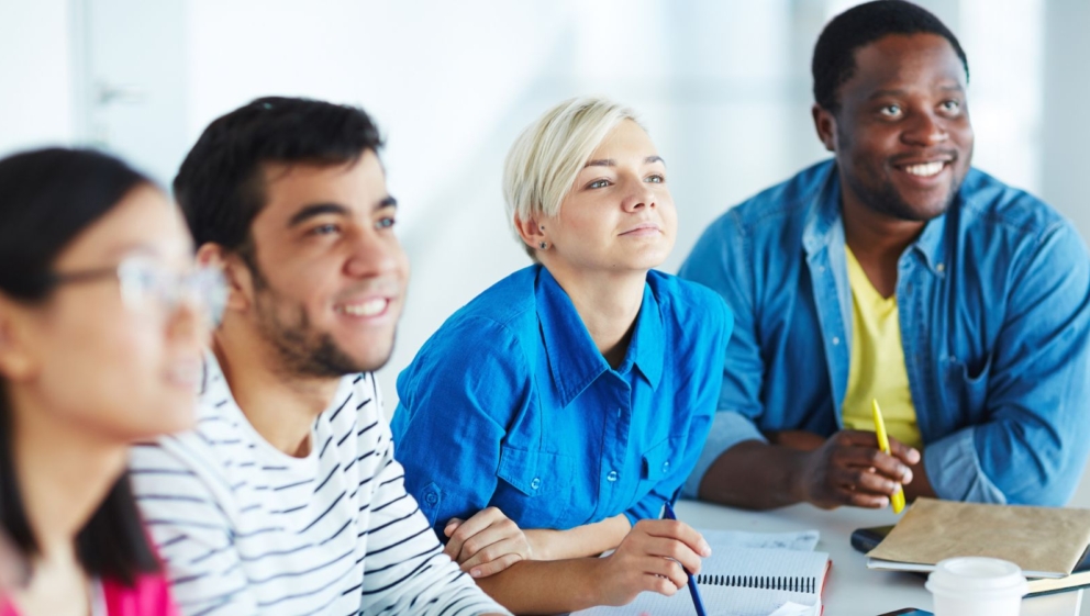Four people, sat smiling as they listen to a session. They have notes, pens and a coffee cup on the table in front of them.