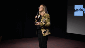 Celia speaking into a microphone on stage, wearing glasses and a camouflage-patterned jacket
