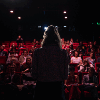 A person standing on stage, viewed from behind, addressing the seated audience in a dimly lit cinema theatre at the premiere
