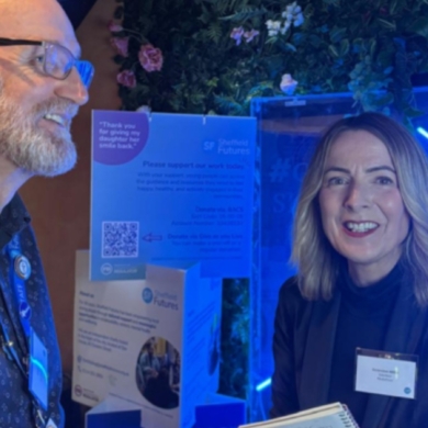 A woman with short blond hair is looking at the camera. She is standing with a man and they are both at a business event