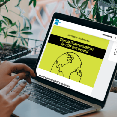 A person is sat in a cafe with plants lining the window. They have a laptop open which shows the Media Trust website page for COP communications and beyond: An online festival.