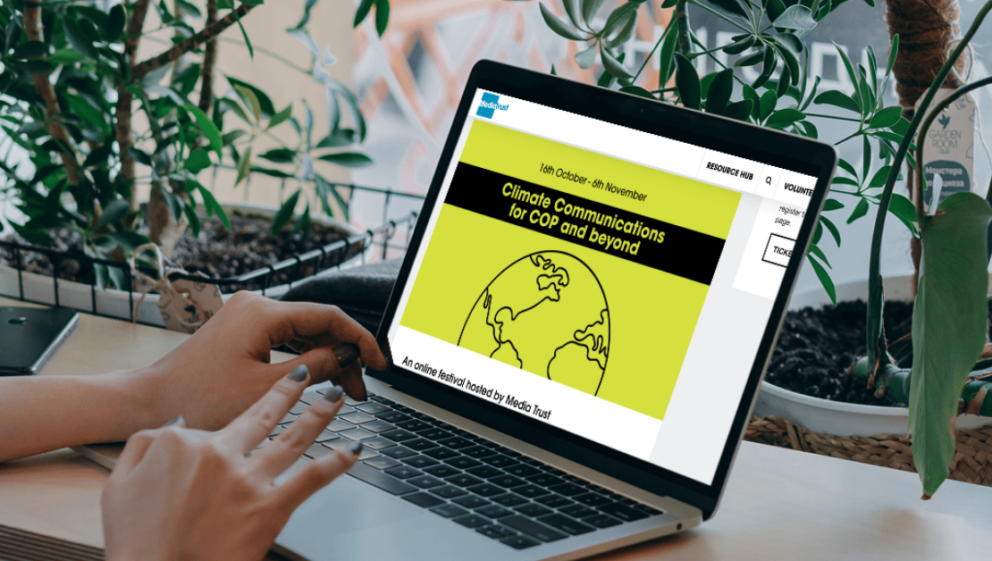 A person is sat in a cafe with plants lining the window. They have a laptop open which shows the Media Trust website page for COP communications and beyond: An online festival.