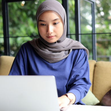 A woman wearing a hijab, sat a desk, working on a laptop.