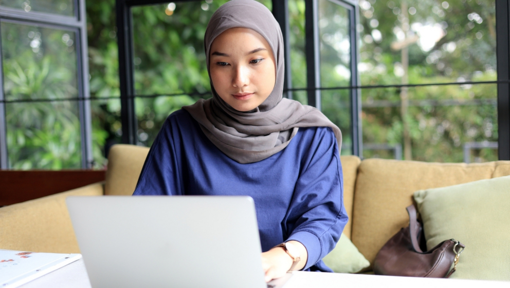A woman wearing a hijab, sat a desk, working on a laptop.