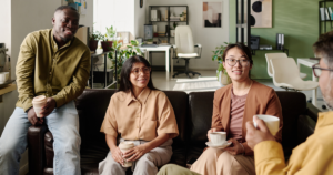 A group of 3 people sit on a workplace sofa with plants in the background, smiling and with mugs in hand. They speak to another person who is only slightly in the shot, also holding a mug.