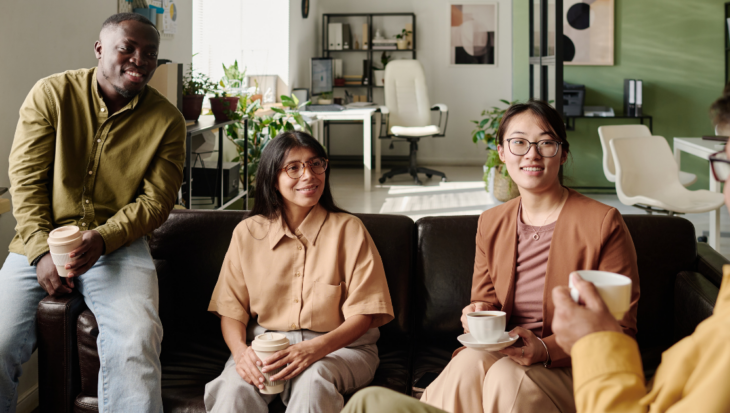 A group of 3 people sit on a workplace sofa with plants in the background, smiling and with mugs in hand. They speak to another person who is only slightly in the shot, also a mug.