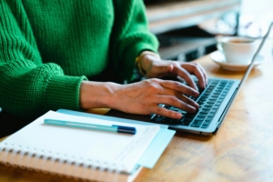 A pair of hands are typing on a grey laptop next to a notepad and pen. The person typing is wearing a bright green jumper.