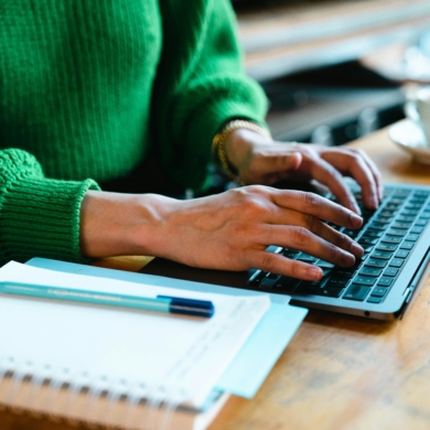 A pair of hands are typing on a grey laptop next to a notepad and pen. The person typing is wearing a bright green jumper.