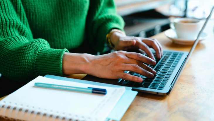A pair of hands are typing on a grey laptop next to a notepad and pen. The person typing is wearing a bright green jumper.