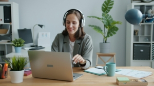A woman with large over head white headphones sits smiling and typing at her laptop. Behind her is a large house plant, and to her side is a notebook and baby blue mug.