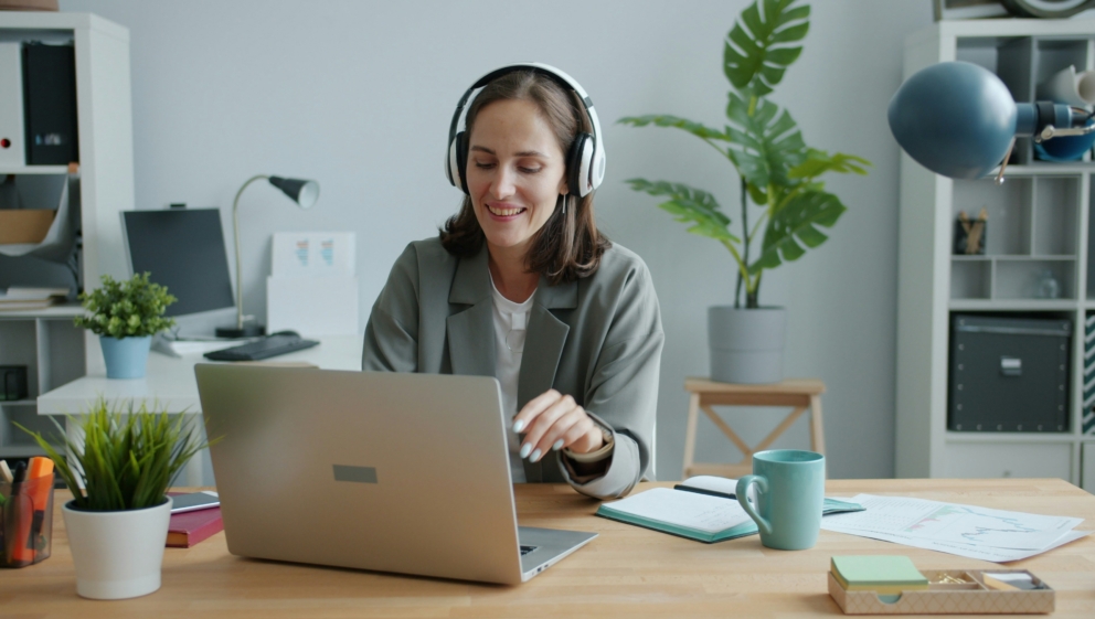 A woman with large over head white headphones sits smiling and typing at her laptop. Behind her is a large house plant, and to her side is a notebook and baby blue mug.